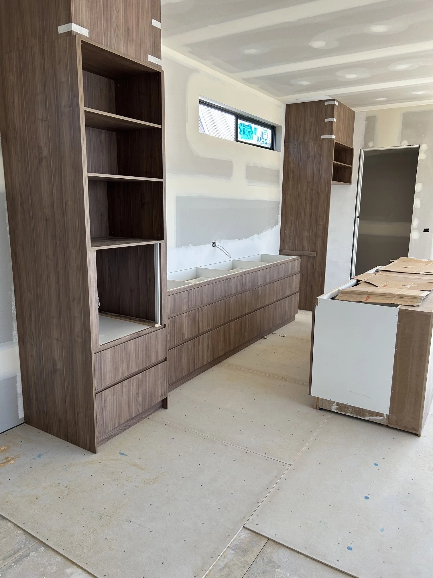 Room under construction with wooden cabinets and shelves, unfinished walls, a small horizontal window, and a concrete floor with paint splatters.