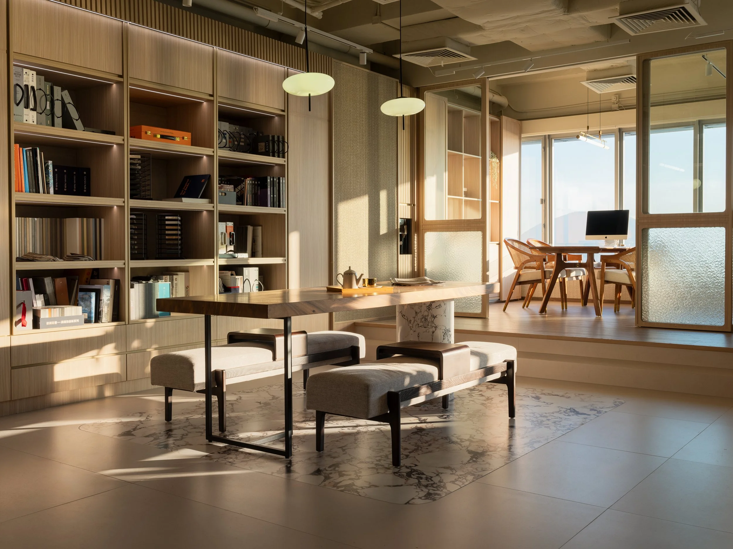 Bright, modern office space with wooden bookshelves, a wooden conference table, and chairs adjacent to a sunlit window. Decor includes pendant lights and a computer on a desk.