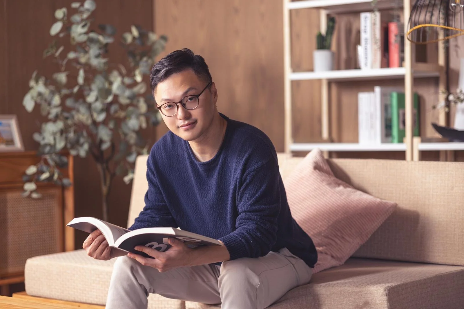 A young man with glasses sitting on a beige sofa, holding an open magazine, in a living room with wooden shelves, books, and a potted plant in the background.