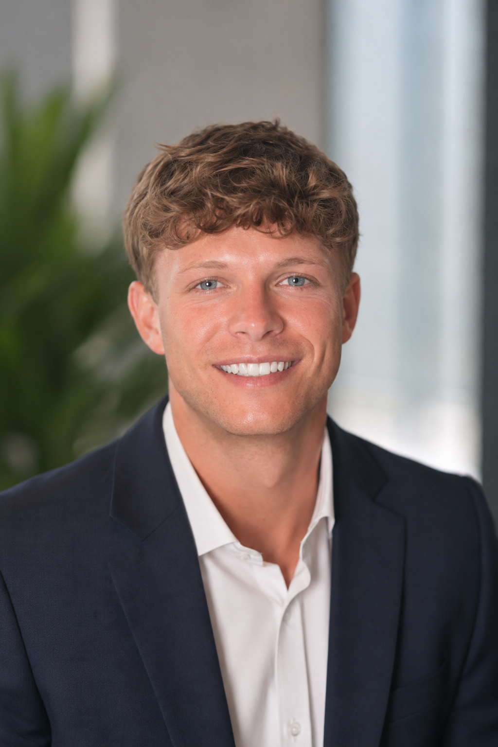 A young man with light skin, wavy light brown hair, and blue eyes, wearing a white shirt and dark blazer, smiling at the camera.
