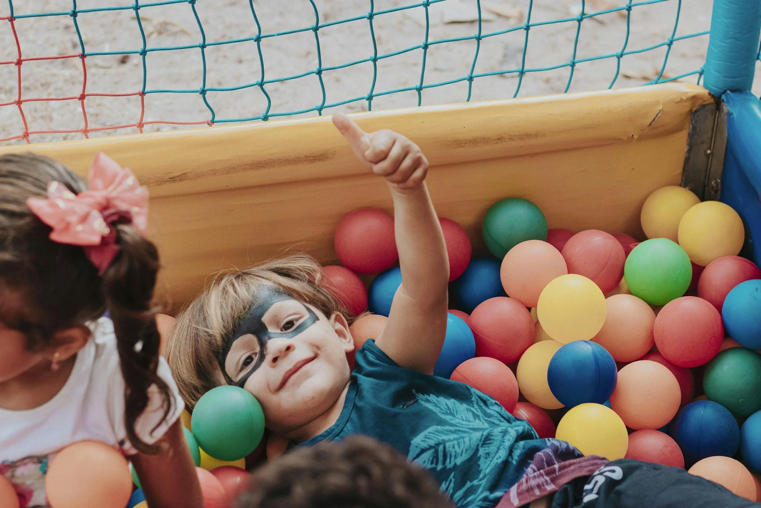 Children playing in a ball pit with colorful plastic balls, some children have face paint and are smiling, and one child is giving a thumbs-up.