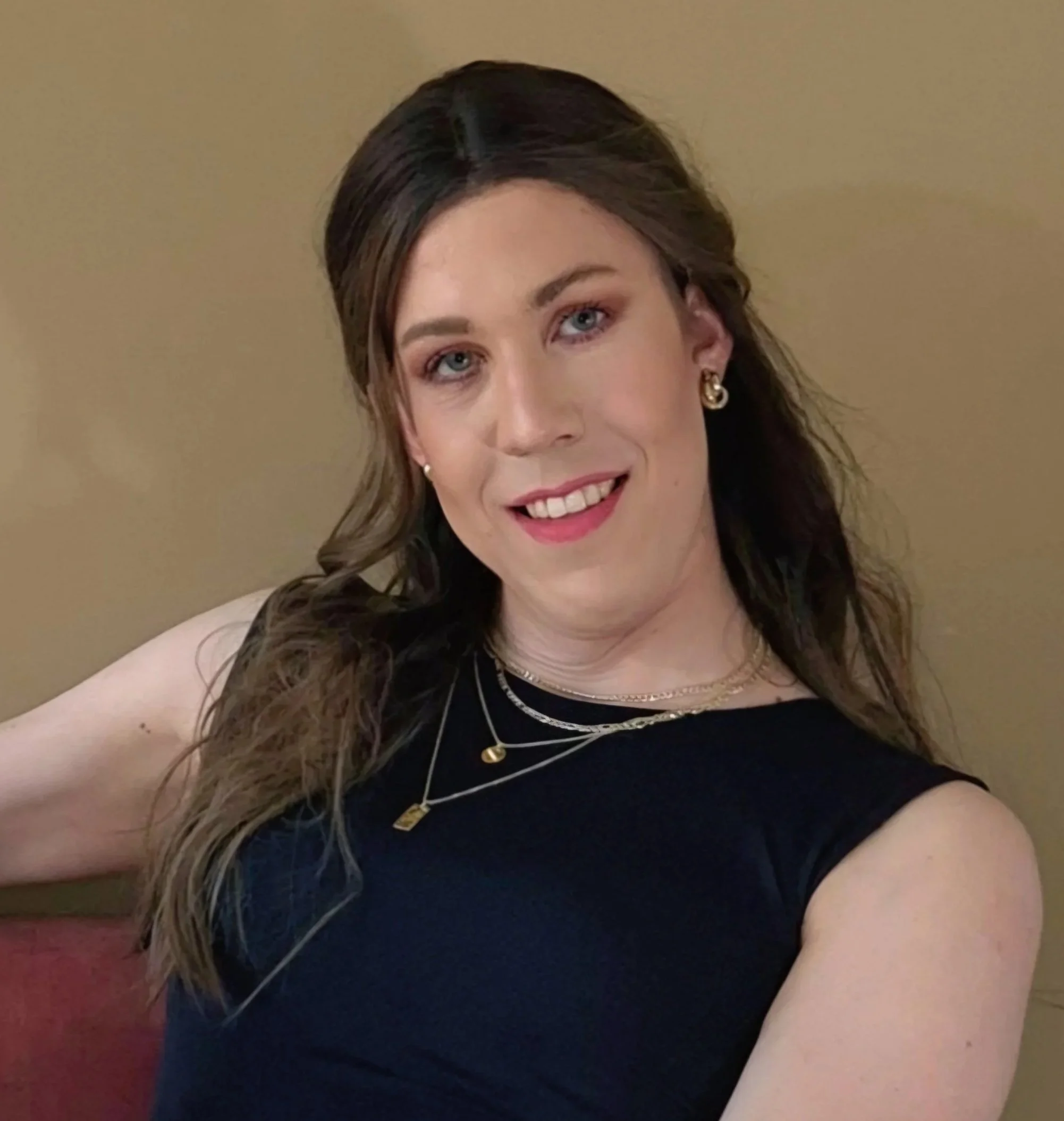 Close-up of Lauren with brown hair and blue eyes wearing gold earrings, layered necklaces, and a black sleeveless top, smiling gently, against a beige wall.