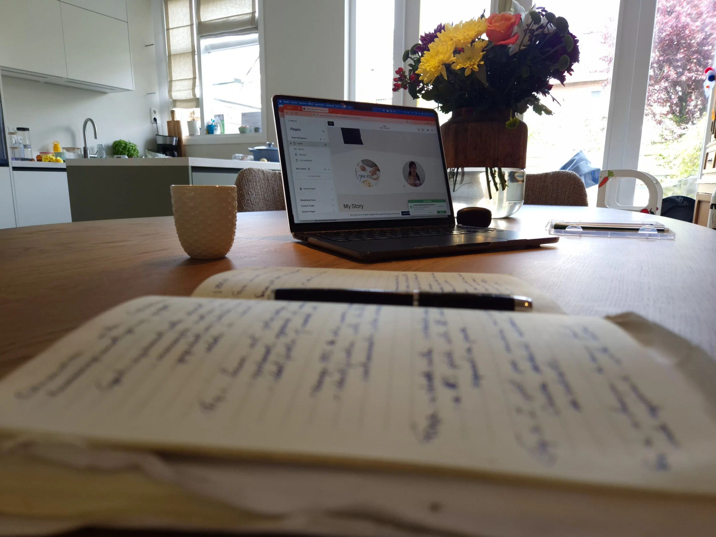 A view of a kitchen and dining area through a window, with a laptop on the table displaying a social media page, a flower vase with colorful flowers, a white mug, and a notebook and pen in the foreground.