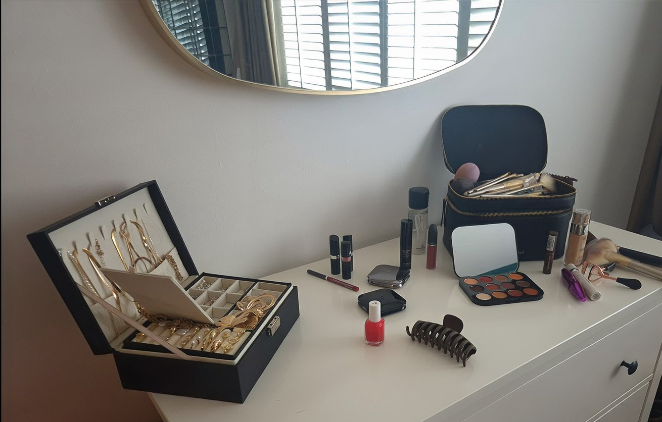 Dresser with jewelry box, makeup, and beauty products on top, mirror reflecting window blinds, and a chair behind the makeup bag.
