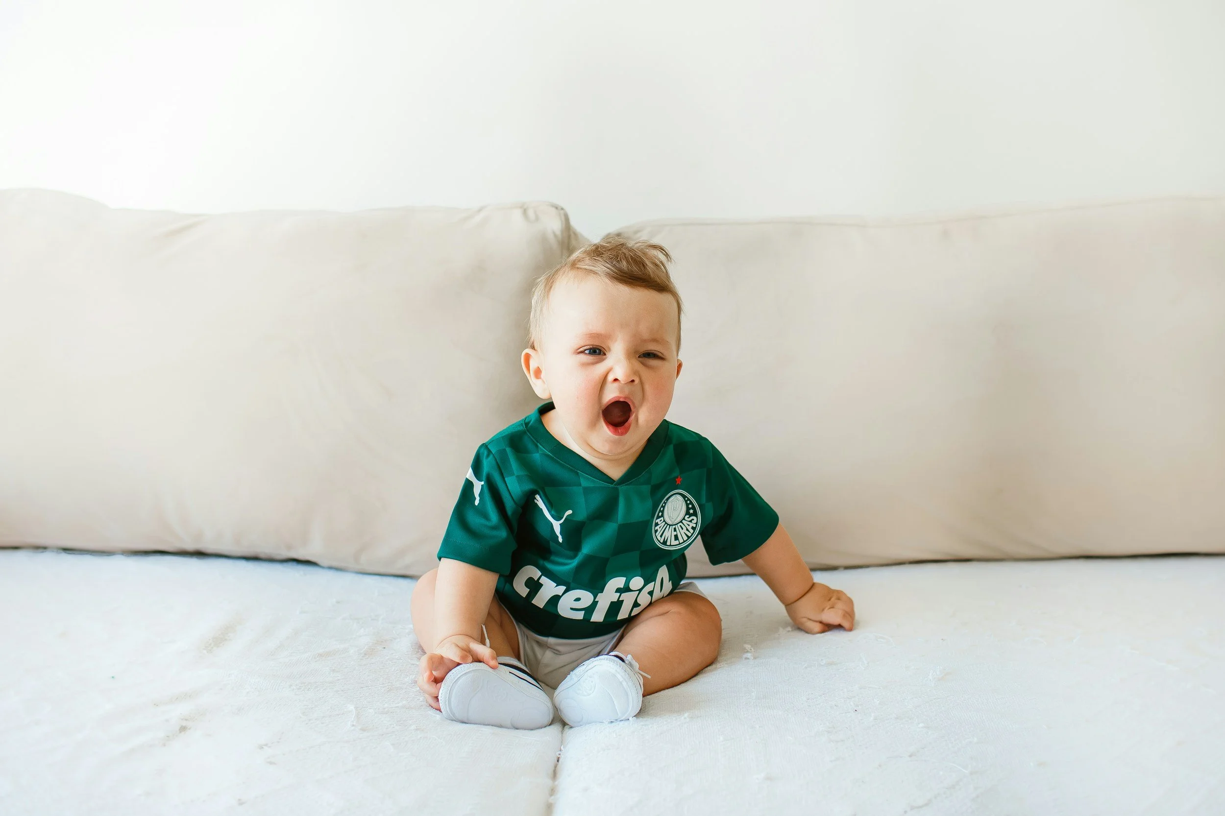 A young child with light hair and blue eyes sitting on a white bed, yawning or shouting, wearing a green soccer jersey with white shoes.