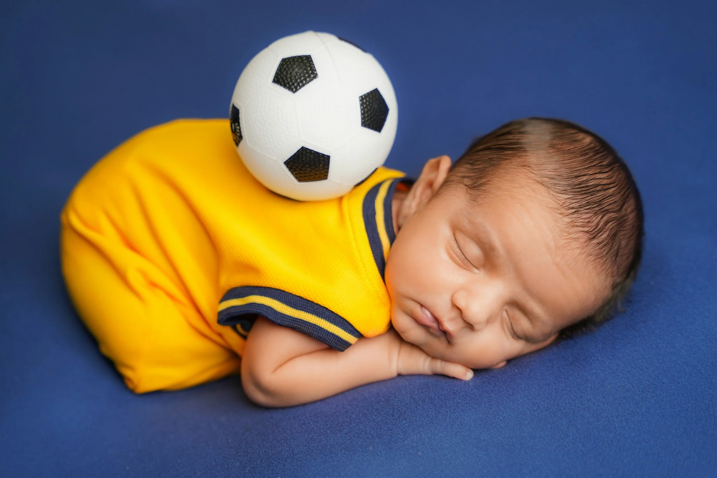 A young boy with short dark hair sleeping on a blue surface with his arms folded under his head. A soccer ball rests on his shoulder.