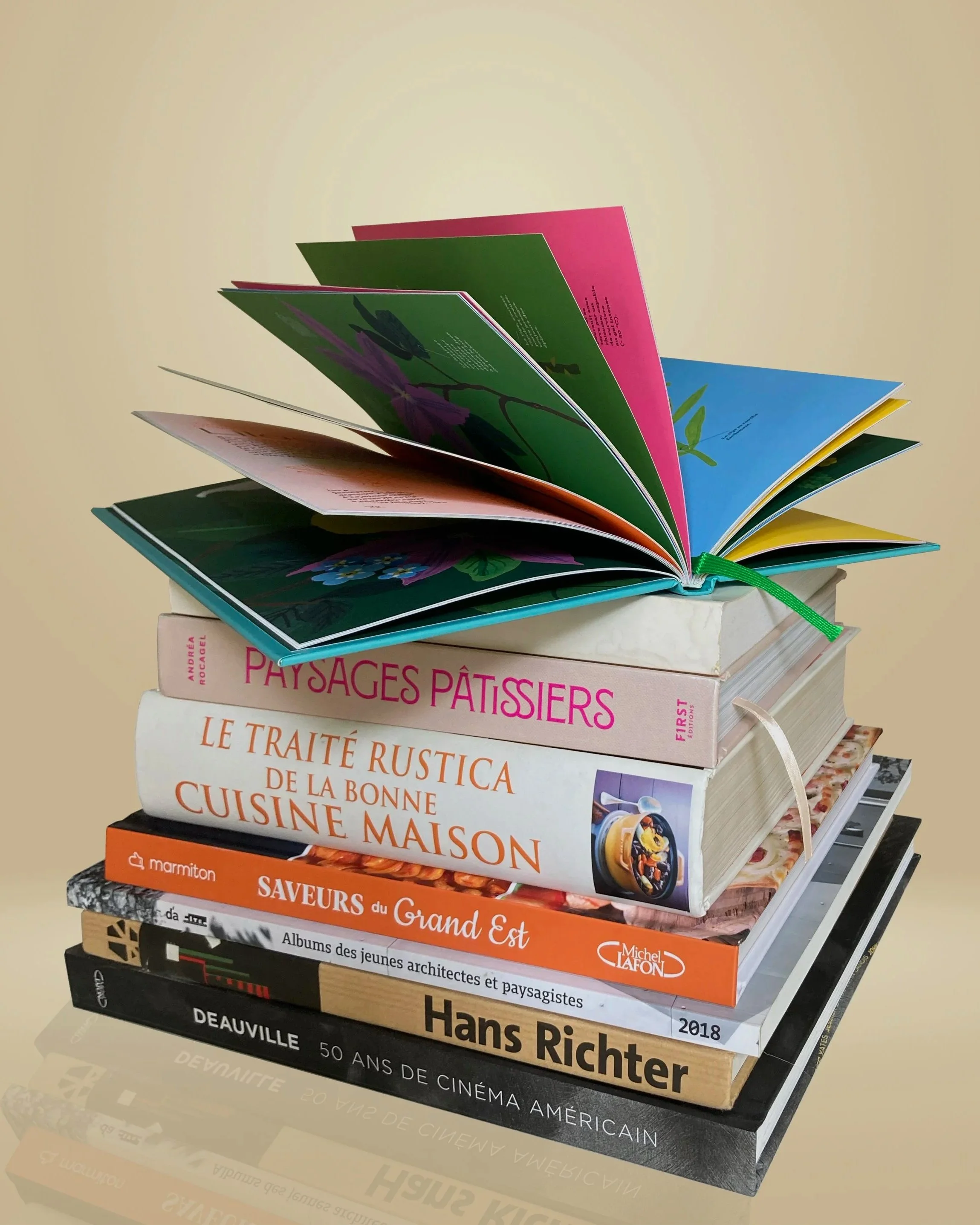 Une pile de livres variés, dont certains portent sur l'art, le design, la cuisine et la culture, avec un livre d'images colorées ouvert en haut.