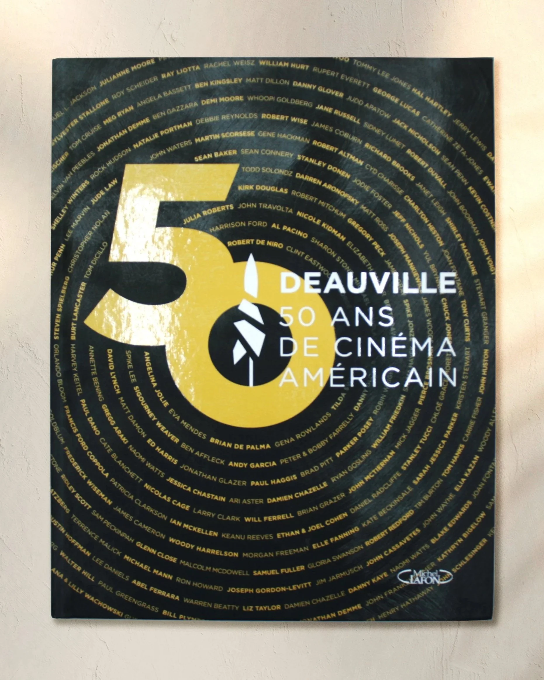 Un livre commémorative en l'honneur des 50 ans du festival du cinéma américain, avec une multitude de noms d'acteurs et réalisateurs célèbres disposés en cercles concentriques. Le texte indique 'Deauville 50 ans de cinéma américain'.