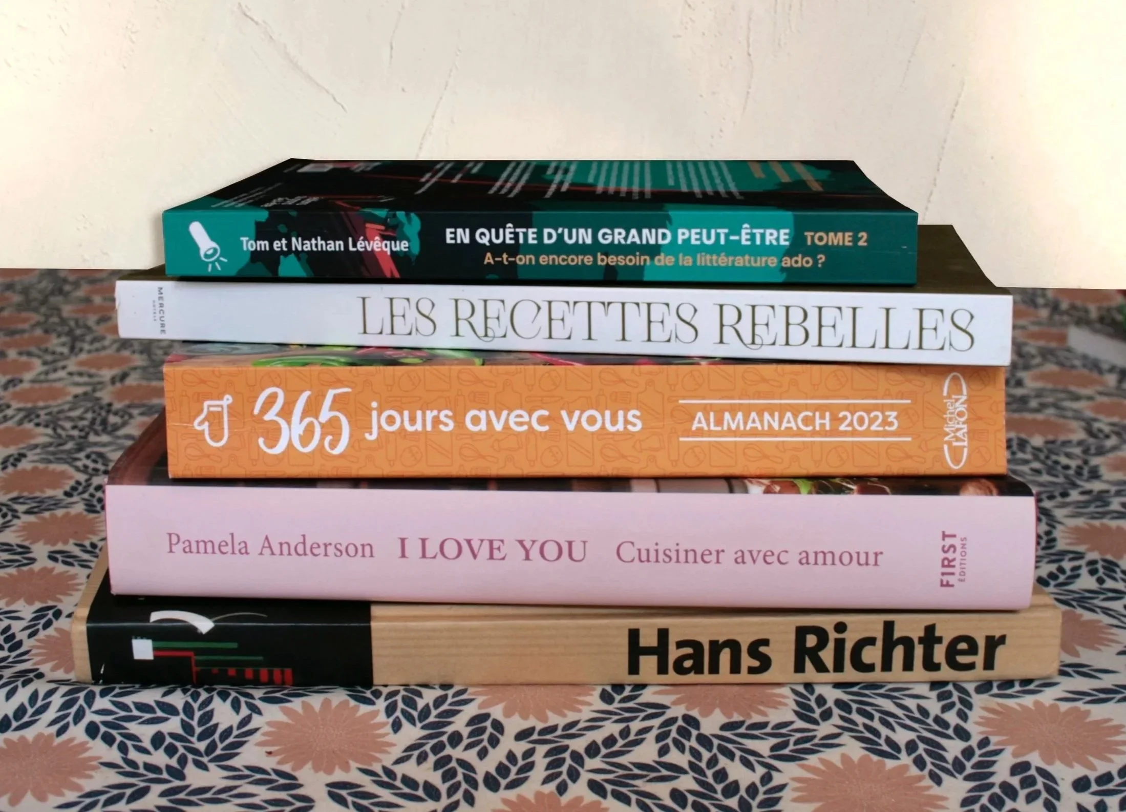 Une pile de cinq livres empilés, avec le sommet reposant contre un mur blanc. Le premier livre en haut est vert, intitulé "En quête d’un grand peut-être, tome 2". Le deuxième livre est blanc, intitulé "Les recettes rebelles". Le troisième est orange, intitulé "365 jours avec vous" de Pamela Anderson. Le quatrième est blanc avec du texte en rouge, intitulé "I love you". Le cinquième livre en bas est noir avec un graphique, signé Hans Richter.