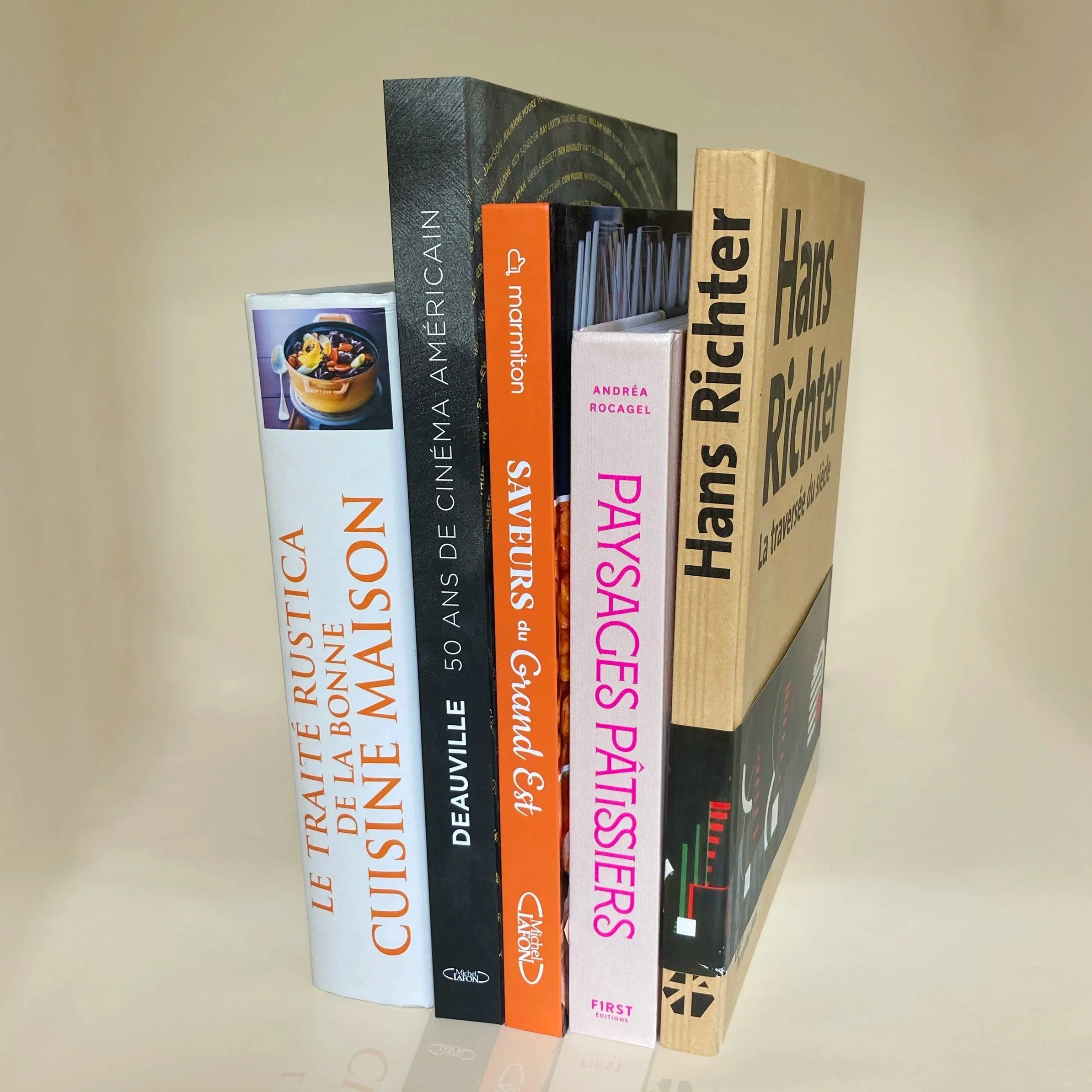 Une pile de plusieurs livres empilés les uns sur les autres. Les titres visibles incluent 'Le plaisir de cuisiner', 'La cuisine de ma mère', 'First Editions' et d'autres. La photo est prise de côté, montrant le côté épais des livres.