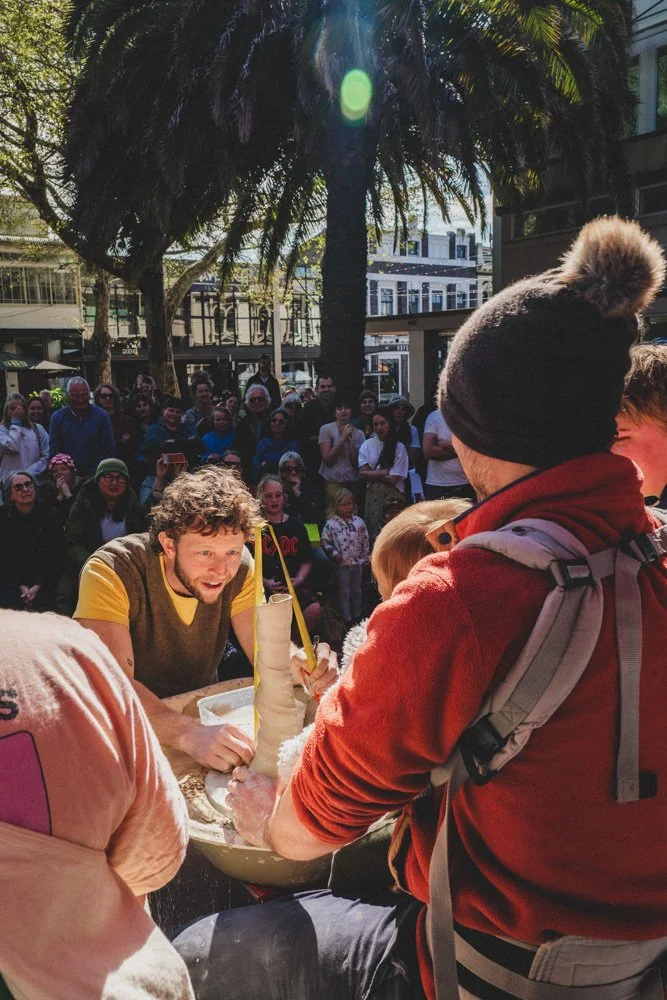 A street performer creating a balloon sculpture while a crowd watches, outdoors in a city with palm trees.