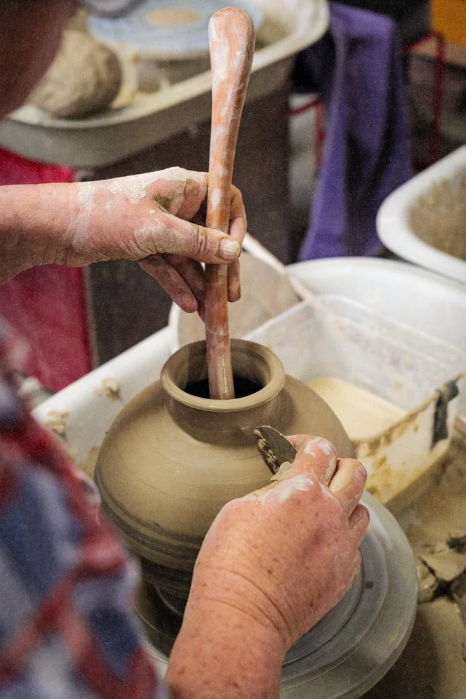 Someone is shaping a clay pot on a pottery wheel using tools, with other pottery materials in the background.