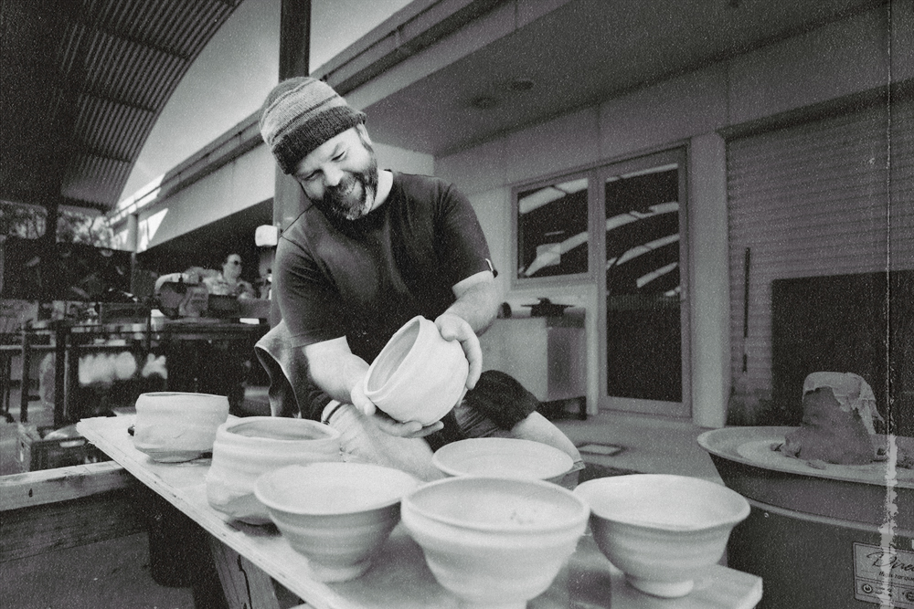 A man with a beard and a beanie, smiling, stacking bowls on a wooden table outdoors, with additional bowls and a hat in the background.
