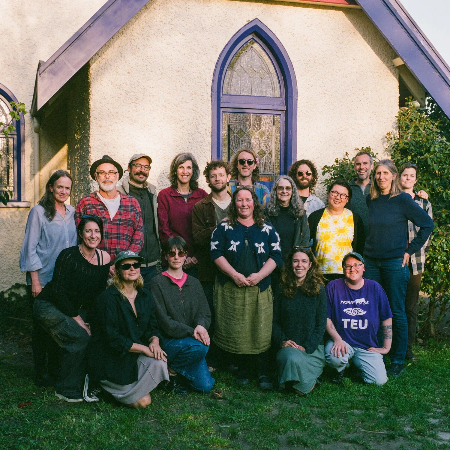Group of 16 people posing outdoors in front of a church with a stained glass window, during daytime.