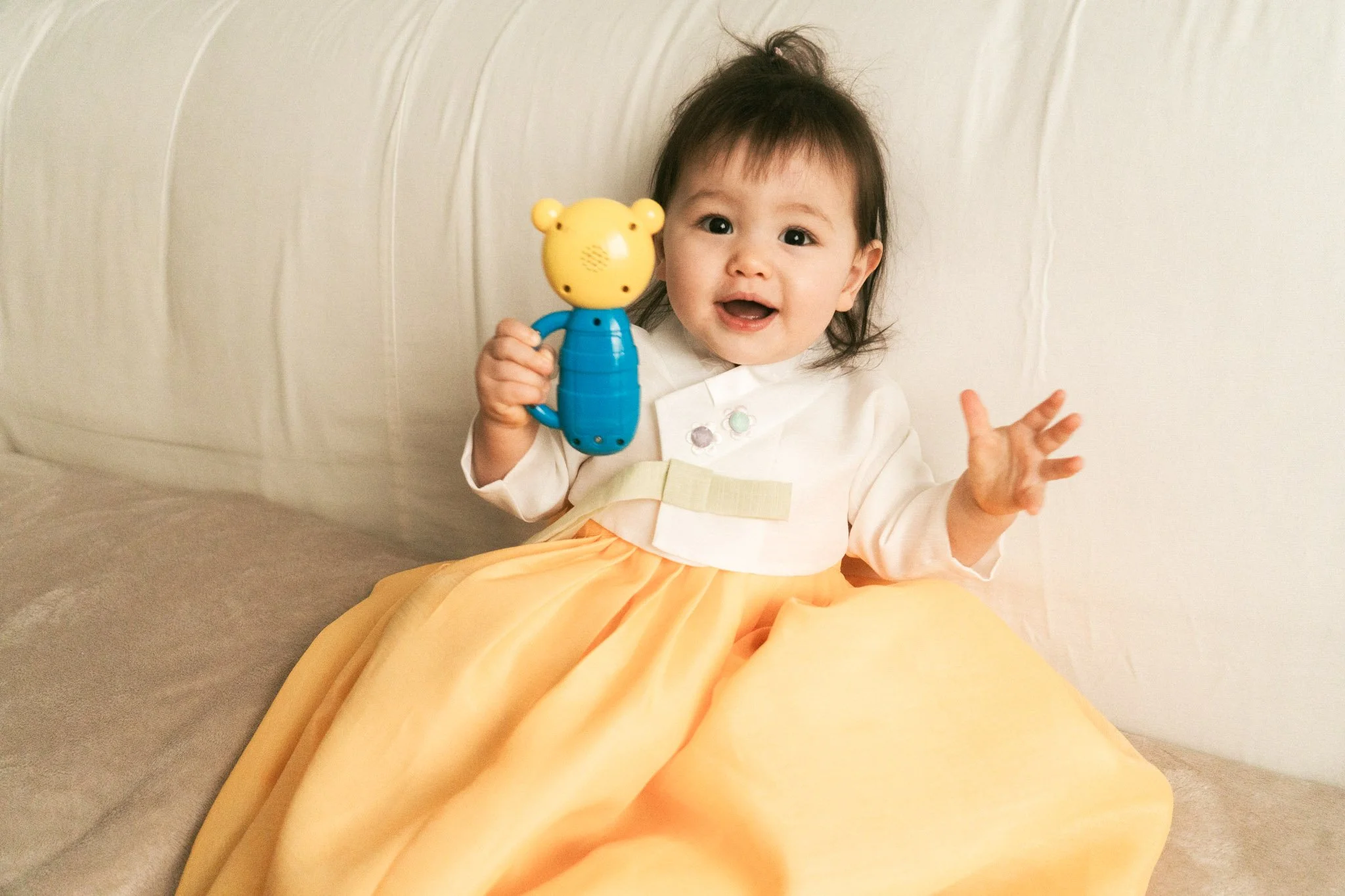 Ein lachendes Baby mit dunklem Haar hält ein Spielzeug in Form eines Teddybärs. Das Baby sitzt auf einem beige-gelben Stoffhintergrund.