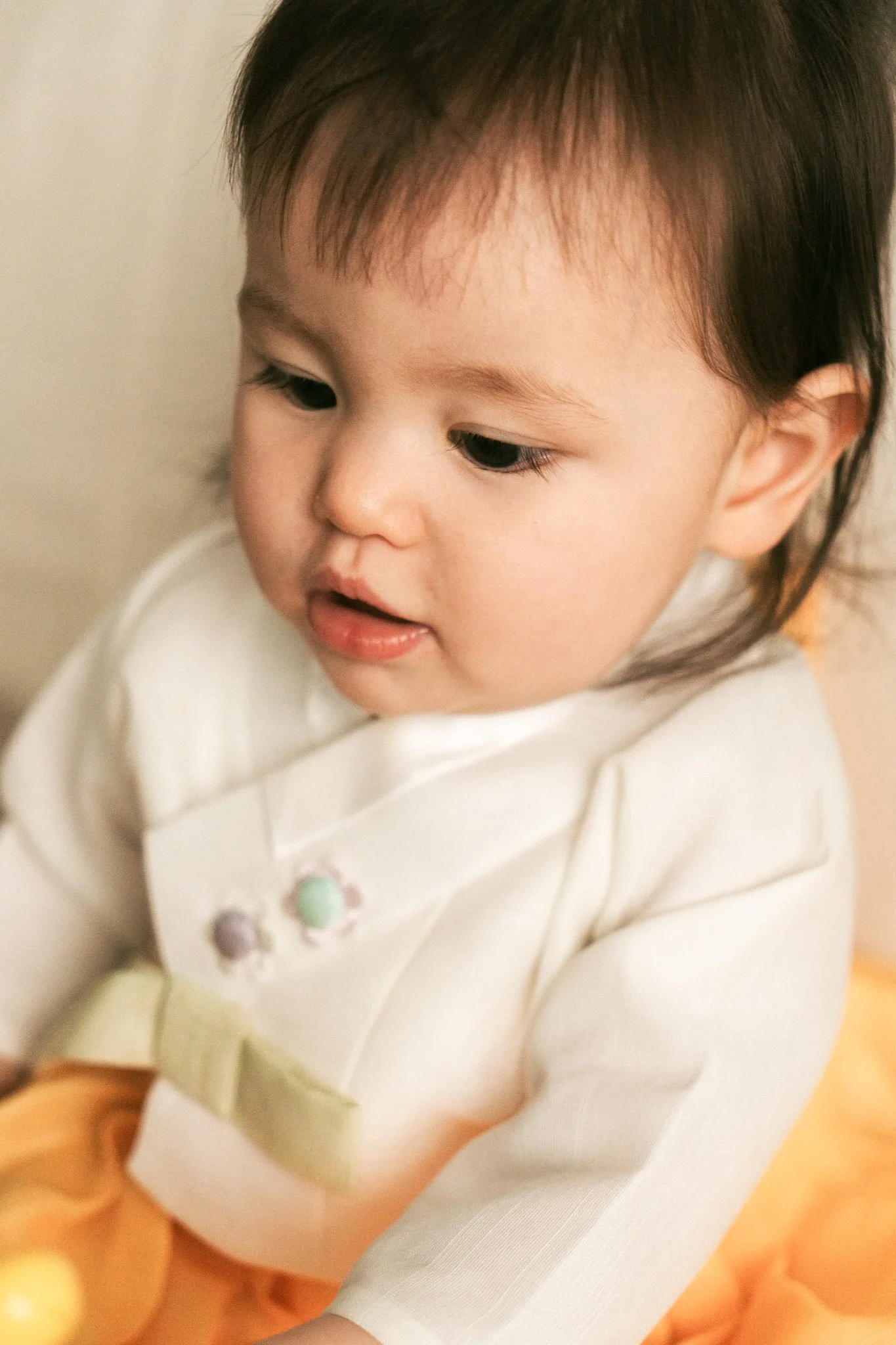 Close-up eines Babys mit dunklen Haaren, das eine weiße Kleidung trägt und auf den Boden schaut.