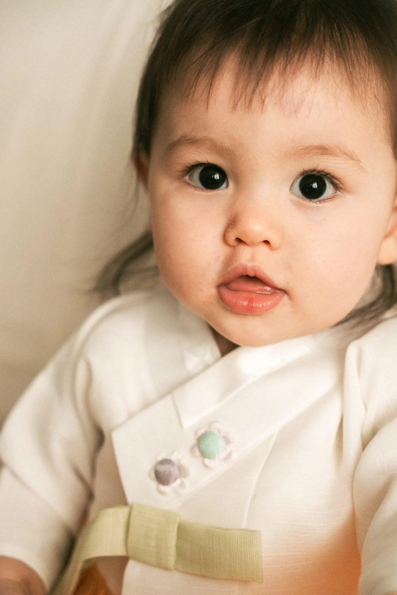 Nahaufnahme eines süßen, kleinen Babys mit großen, dunklen Augen und offenen Lippen, das in einem weißen Outfit sitzt, auf einem Holzboden, mit einem hellen Hintergrund.
