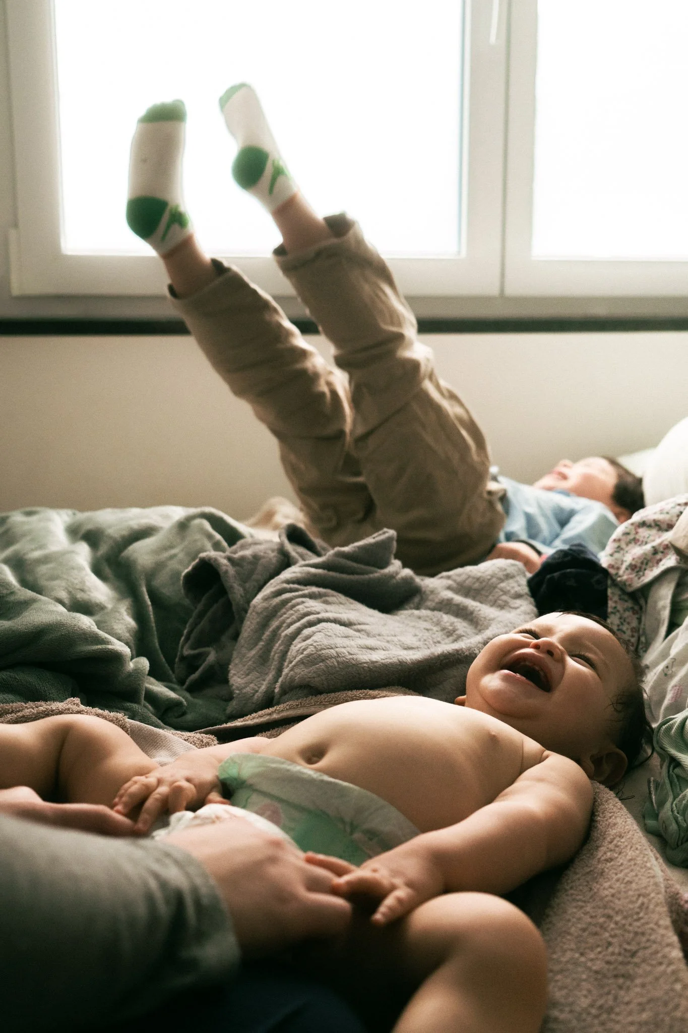 Zwei Kinder, eines braungekleideter Junge und eines blondes Mädchen, lachen und spielen auf einem Bett mit Wolldecken, während sie Polyfilling-Krümel auf ihrem Kopf tragen. Das Mädchen liegt auf dem Rücken und der Junge steht auf den Beinen, hebt die Beine in die Luft, vor einem Fenster mit hell erleuchtendem Hintergrund.