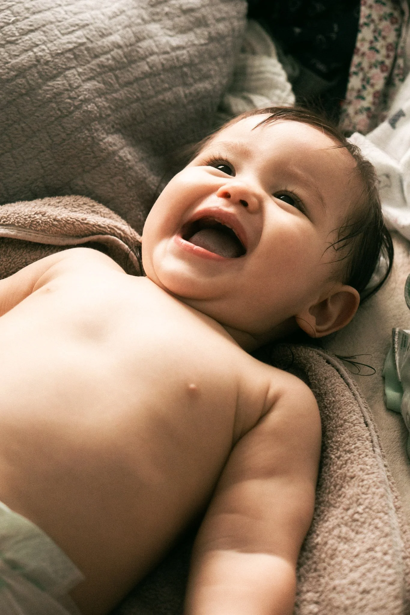 Lächelnder Baby mit dunklen Haaren liegt auf einer beige Stoffdecke.