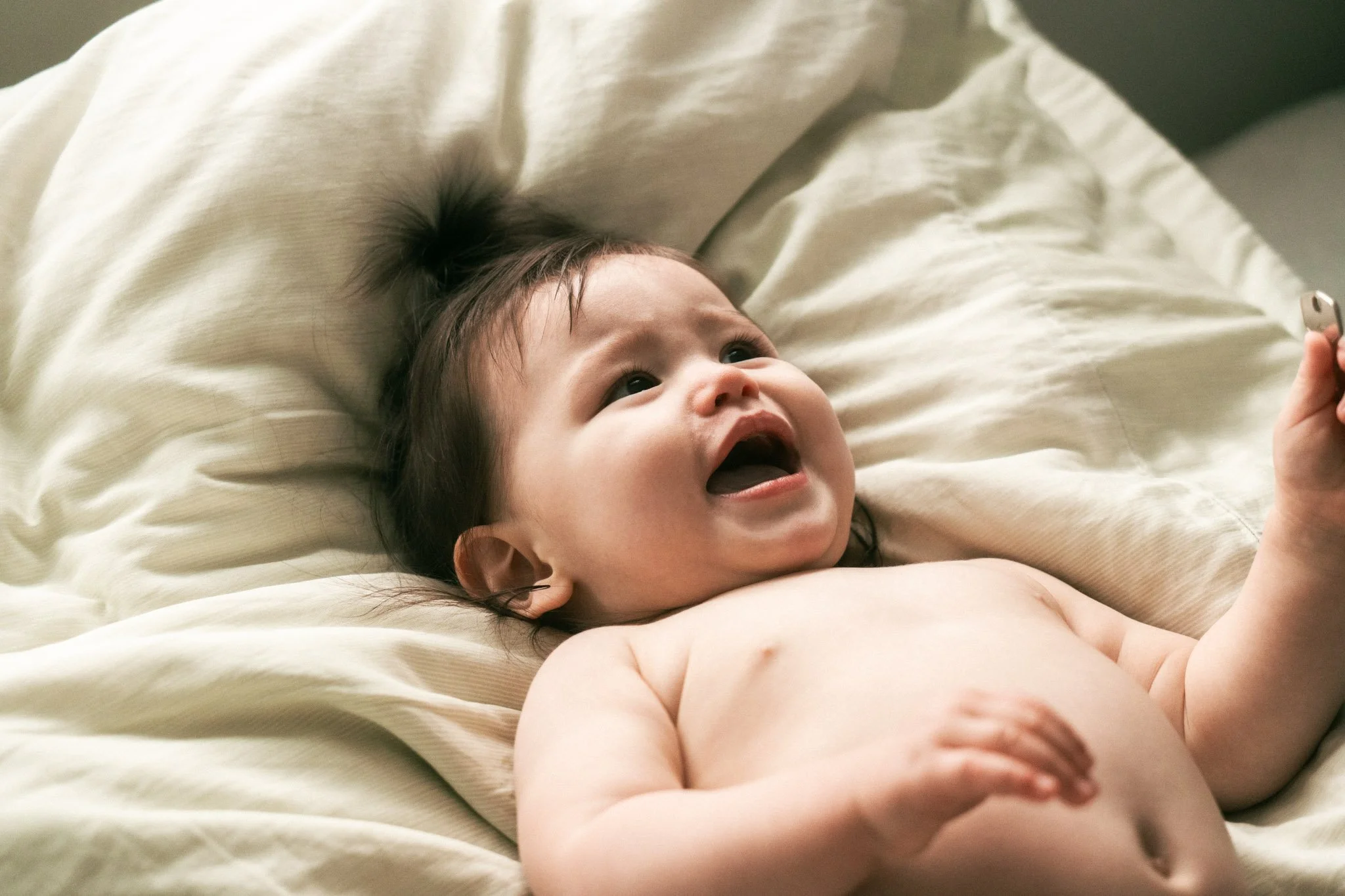 Ein Baby liegt lachend auf einem Bett, umgeben von weißen Bettwäsche, und schaut in die Kamera.