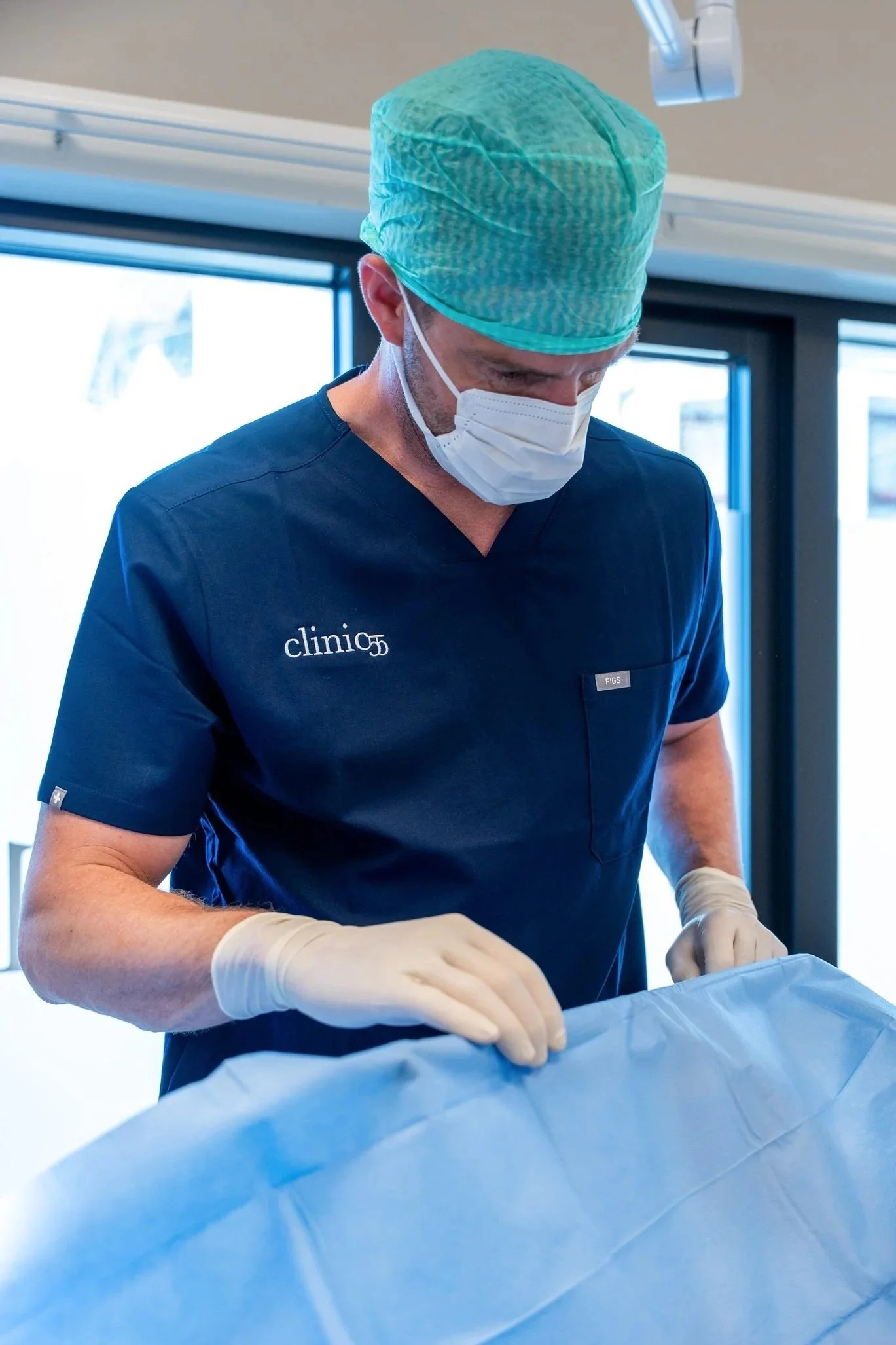 En lege i blå uniform og grønn kirurgisk hatt jobber med medisinske prosedyrer i et operasjonsrom.