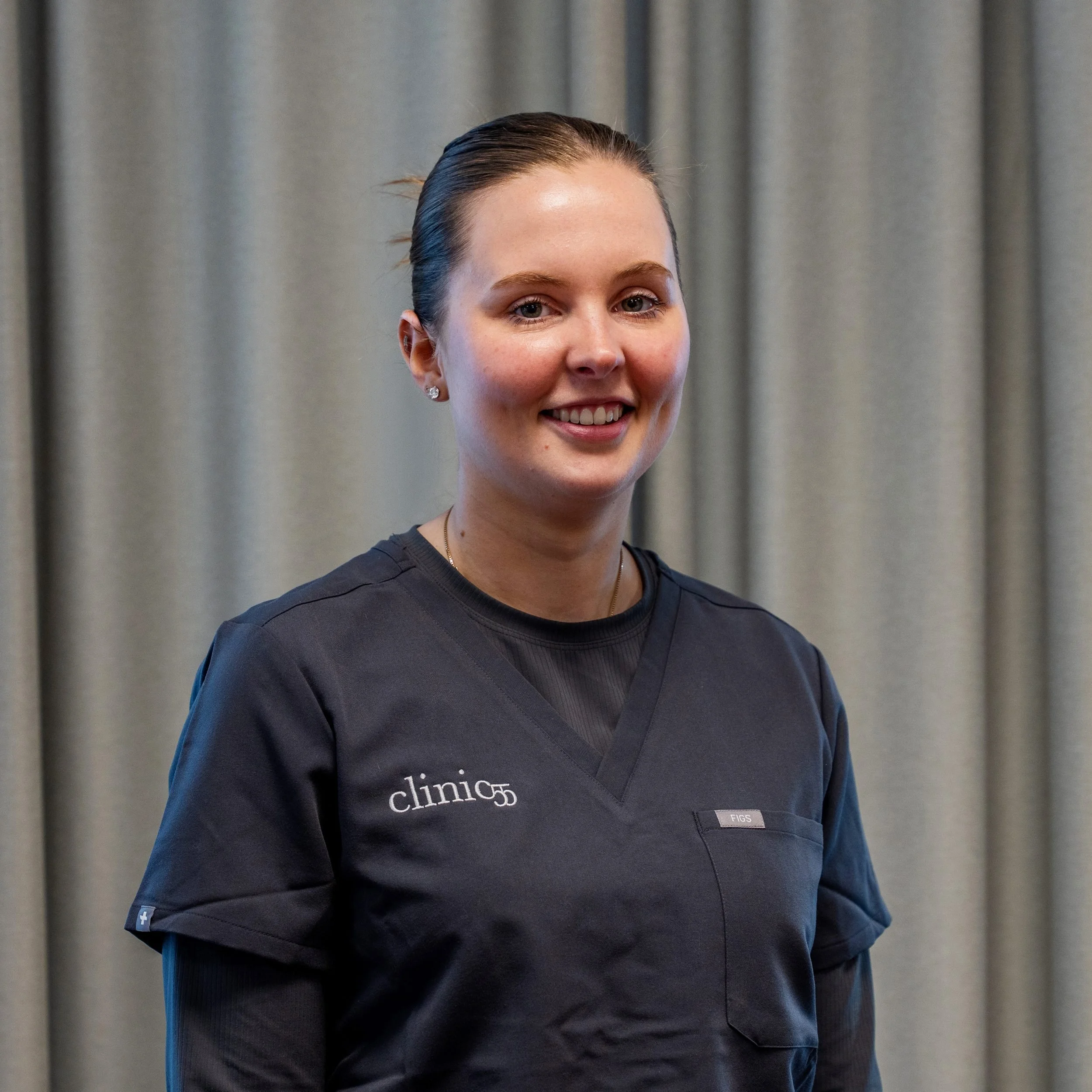 Et bildet av en ung kvinne med mørkt hår, smilende, iført en Klinikkuniform fra clinic 55. står foran en grå gardin.