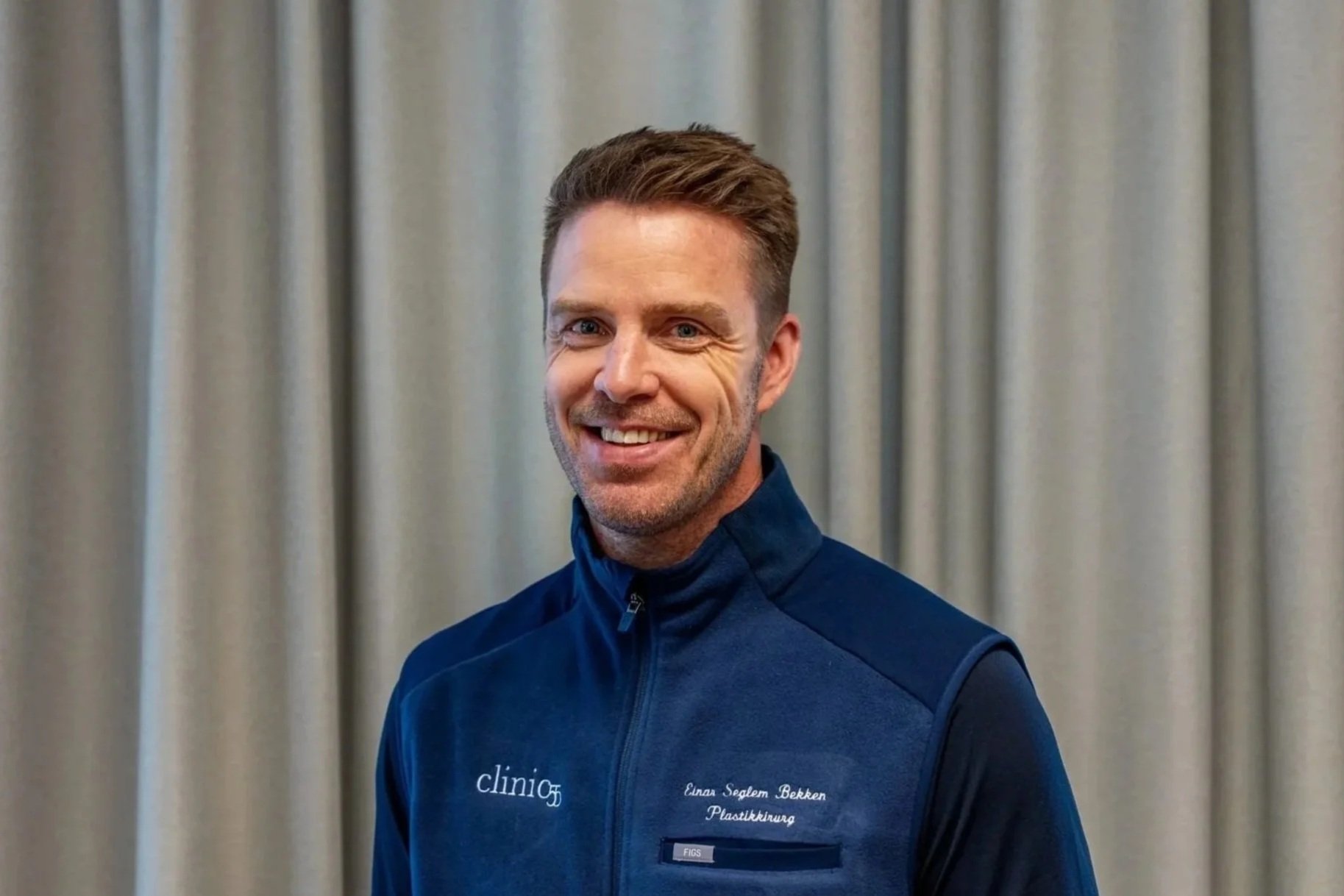 En mann smiler for kamera, iført en blå treningjakke med tekst som sier 'Einar Seglem Bekken Plastikkirurg' og står foran en grå gardin.