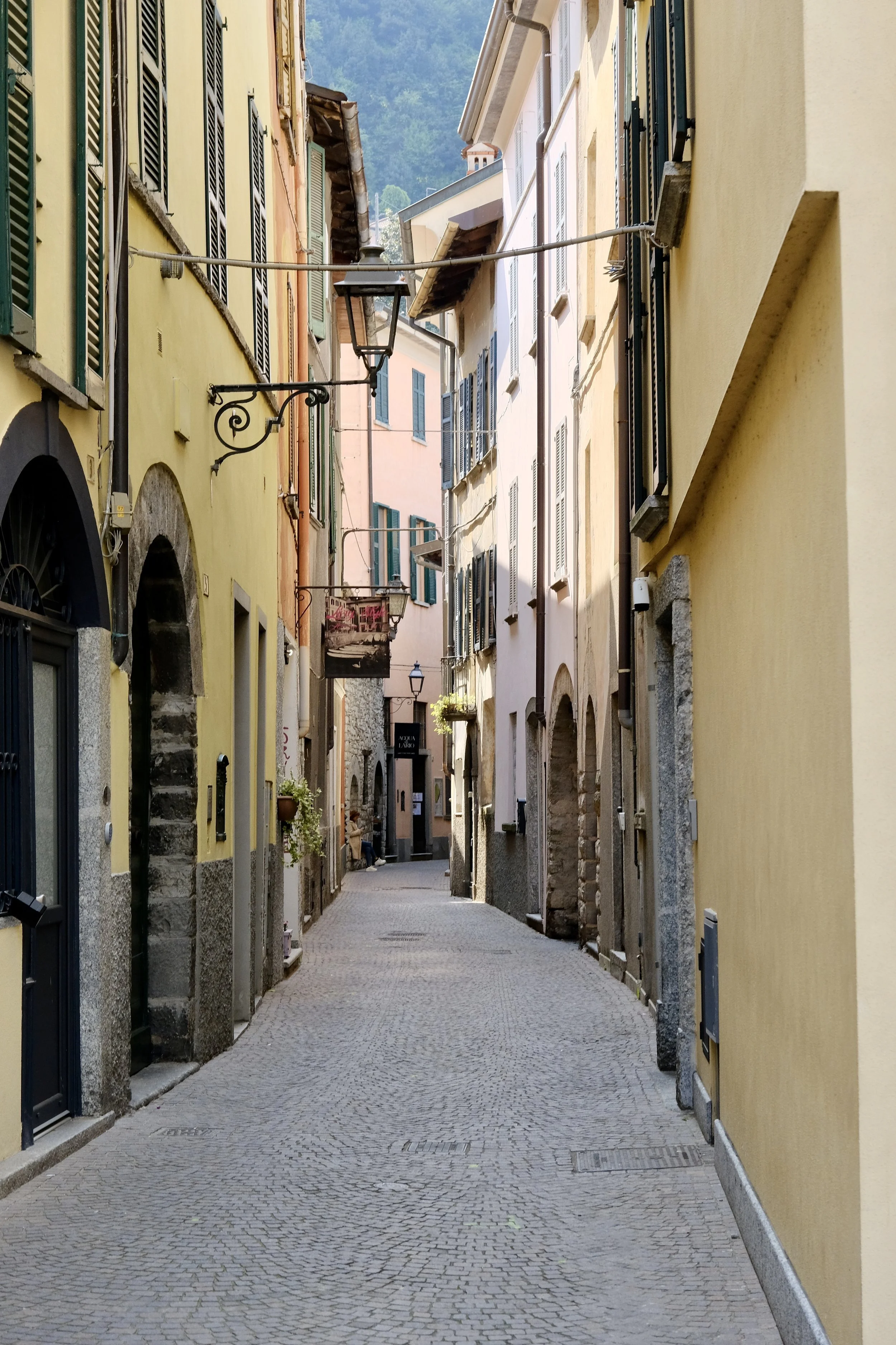 A narrow European street with colorful buildings on both sides, cobblestone pavement, and hanging signs and lamps.