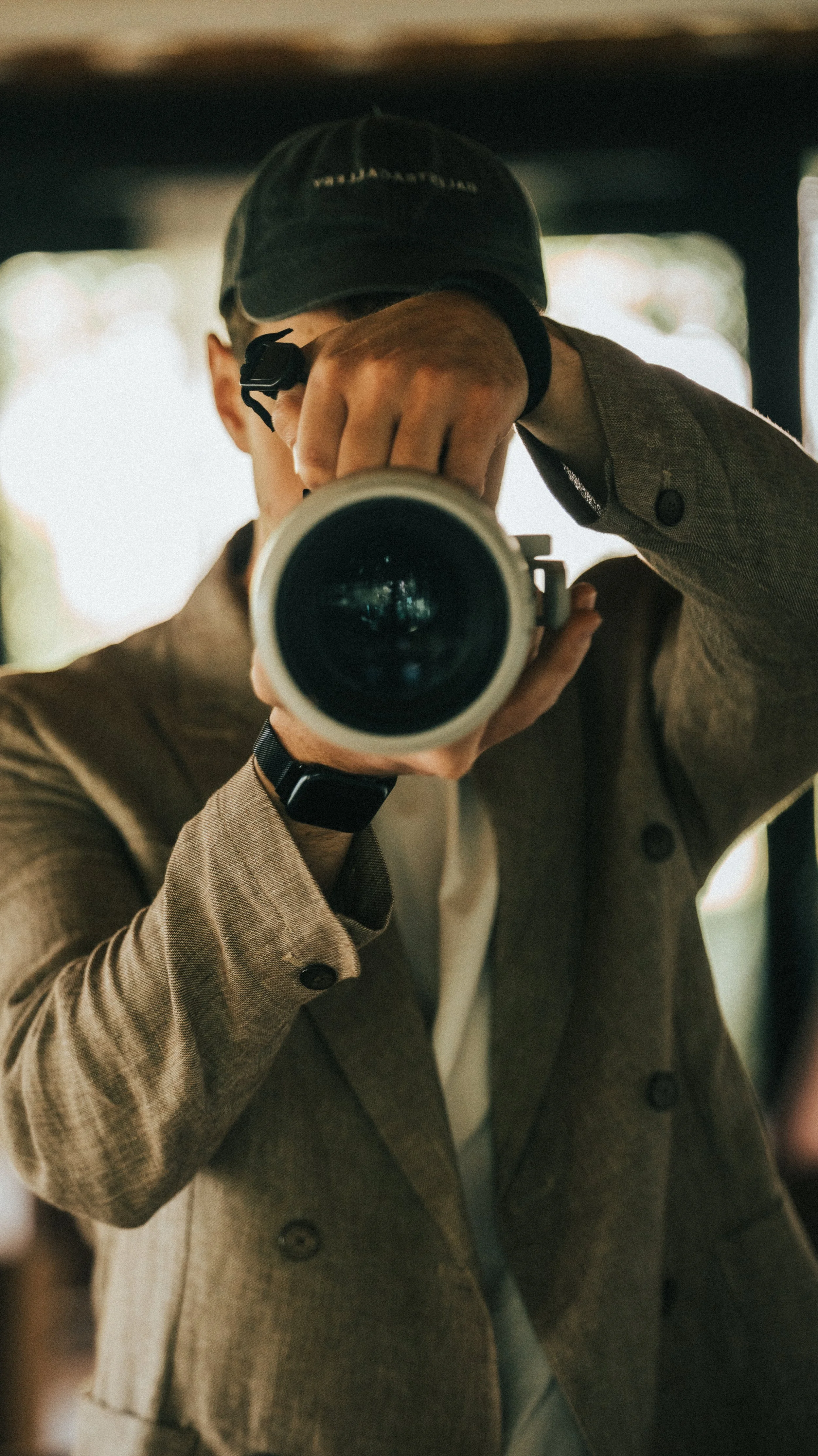 Fotograf eines Mannes in einem braunen Sakko, der in einem Spiegel ein Selfie macht. Er trägt eine schwarze Baseballkappe und eine Smartwatch.
