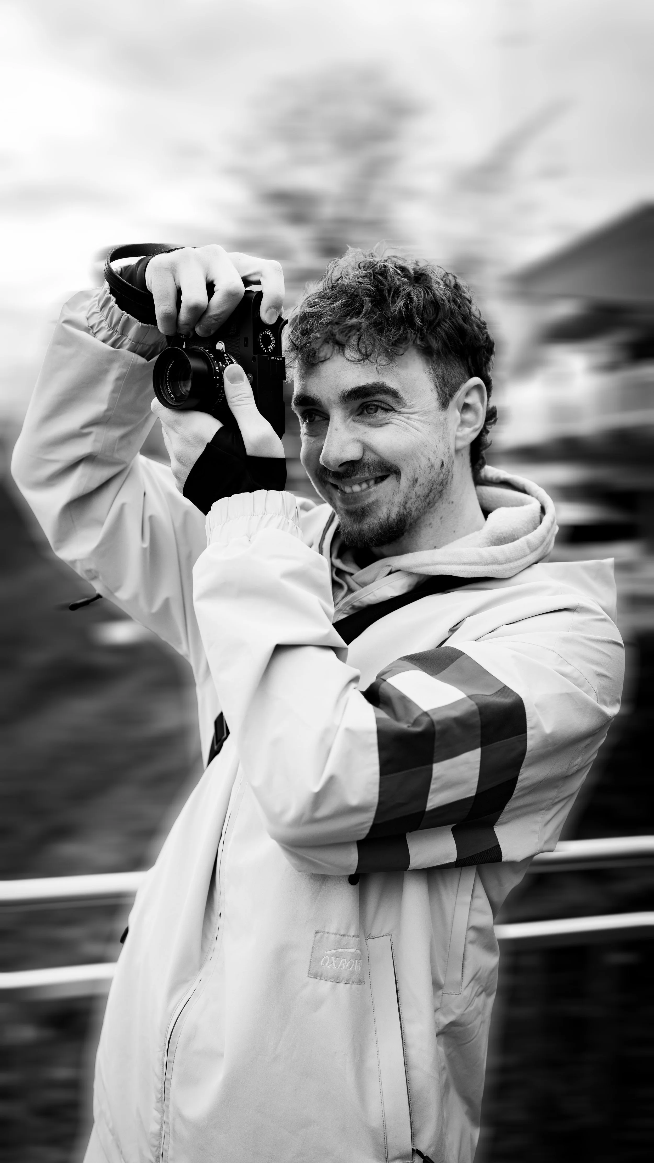 Ein lachender junger Mann mit lockigem Haar fotografiert mit einer Kamera, trägt einen hellen Mantel mit schwarzen und weißen Streifen an den Ärmeln, im Hintergrund ist Wasser und eine bergige Landschaft zu sehen.