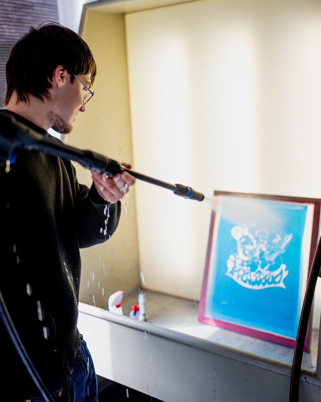 Junger Mann mit Brille reinigt ein Fenster mit einem Hochdruckreiniger, Wasser läuft herunter, im Hintergrund ein aufgehängtes Kunstwerk mit blauer Hintergrundfarbe.
