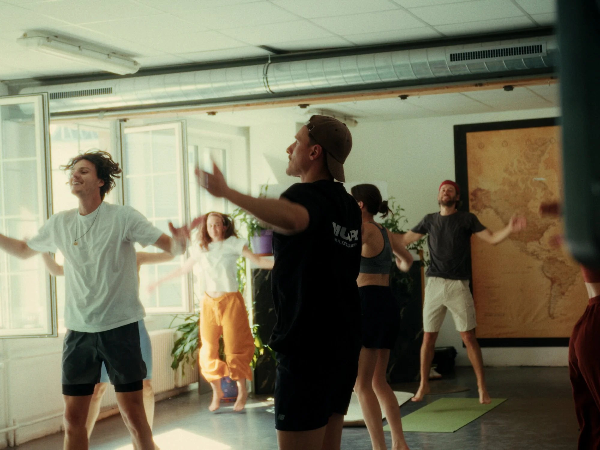 Menschen tanzen in einem Raum während eines Yoga- oder Tanzkurses, mit großen Fenstern im Hintergrund und Pflanzen im Raum.