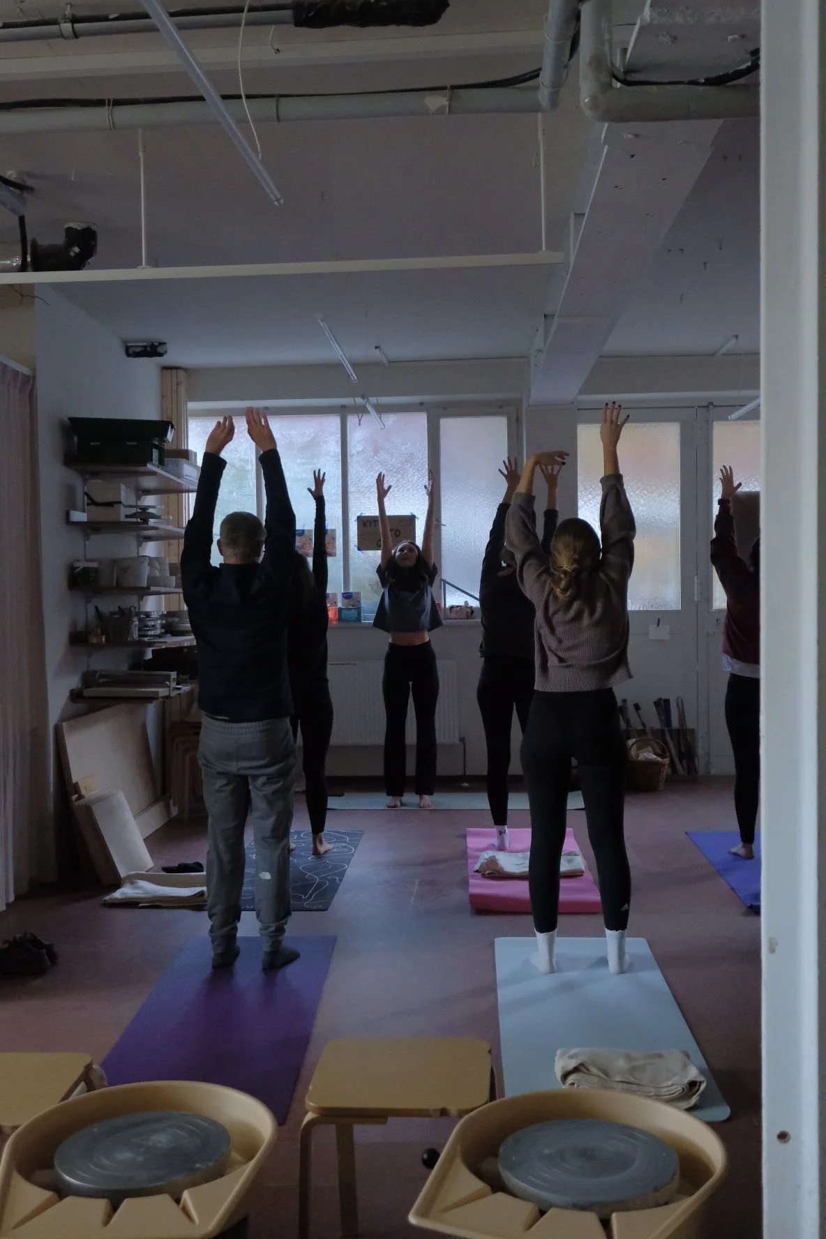 Menschen machen eine Yoga-Klasse in einem Raum mit großen Fenstern, auf Yogamatten, geführt von einer Lehrerin.
