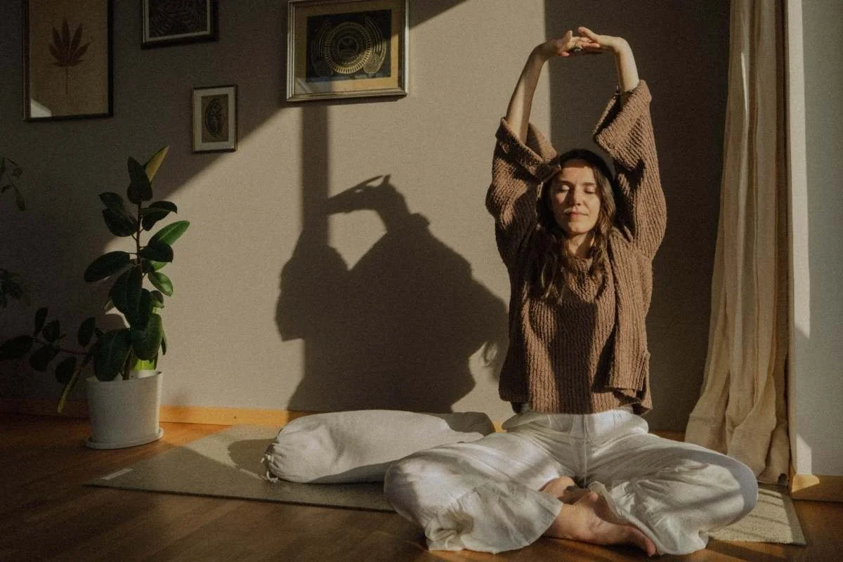 Frau sitzt im Schneidersitz auf einem Teppich, hebt die Arme über den Kopf und macht Dehnübungen, Schatten an der Wand, Zimmer mit Pflanzen und Wandkunst.