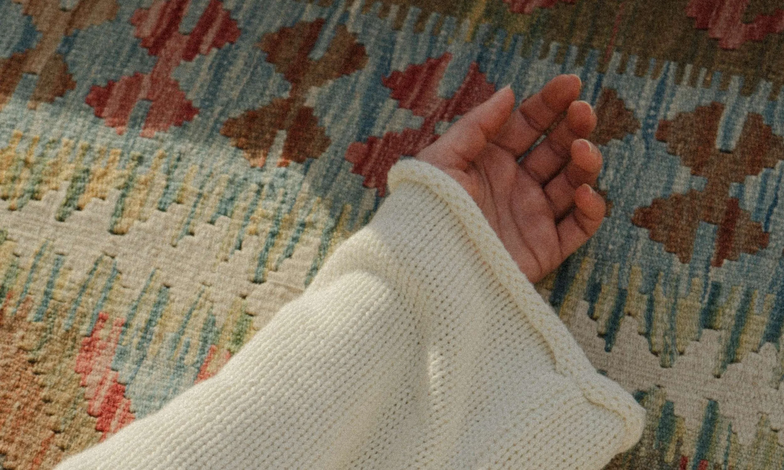 Ein Handteil mit mehreren Ringen, die an den Fingern getragen werden, hält einen weißen Pullover, während der Hintergrund ein bunten, geometrischen Musterteppich zeigt.