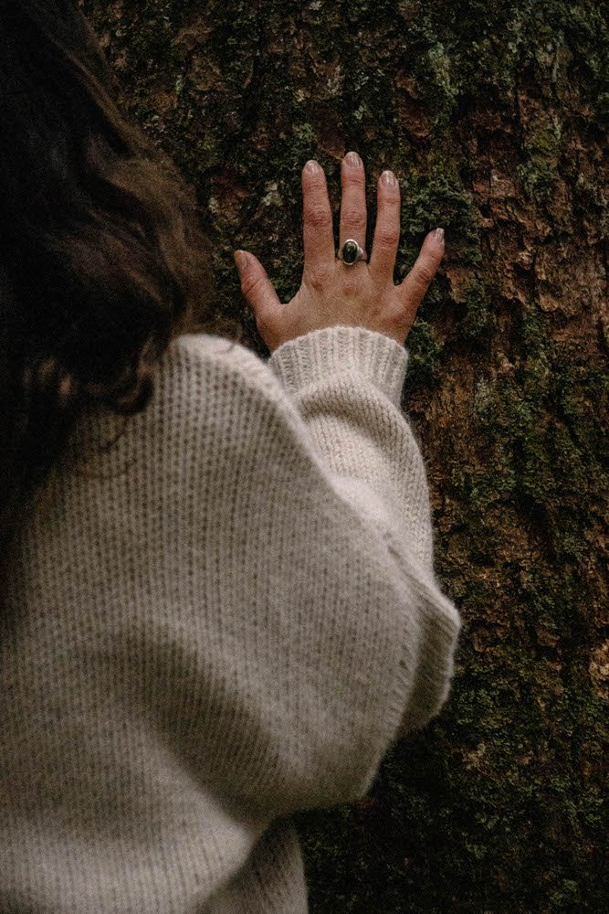 Person berührt Baumrinde mit der linken Hand, trägt einen weißen Pullover, dunkle Haare, Baum mit grober, rissiger Oberfläche.