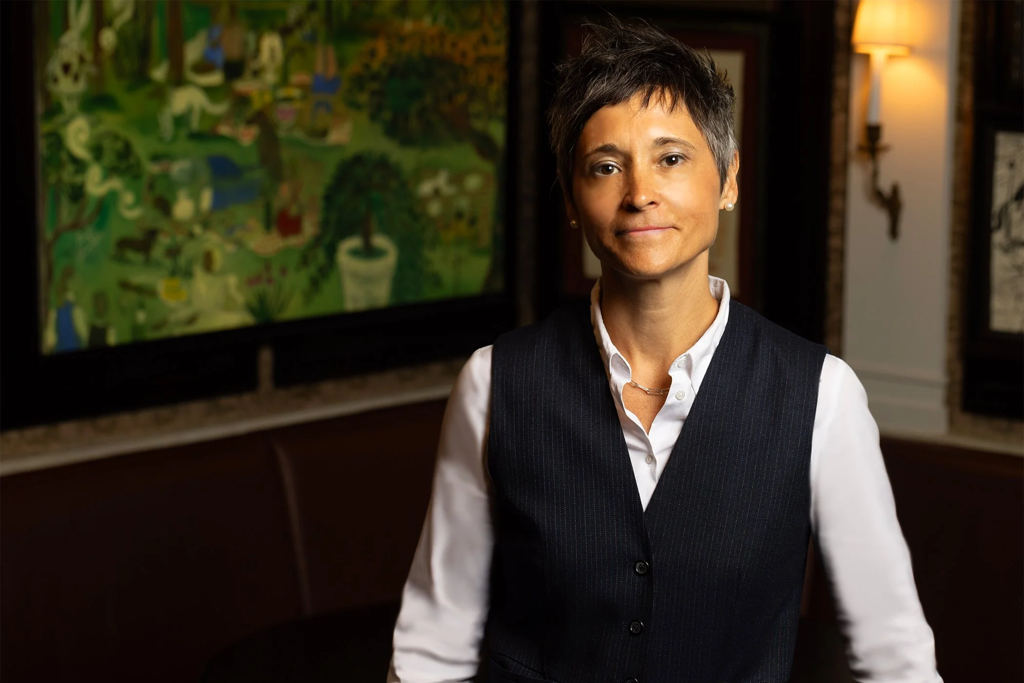 A woman with short gray hair and pearl earrings, wearing a white shirt and a dark pinstripe vest, standing indoors with artwork and warm lighting in the background.