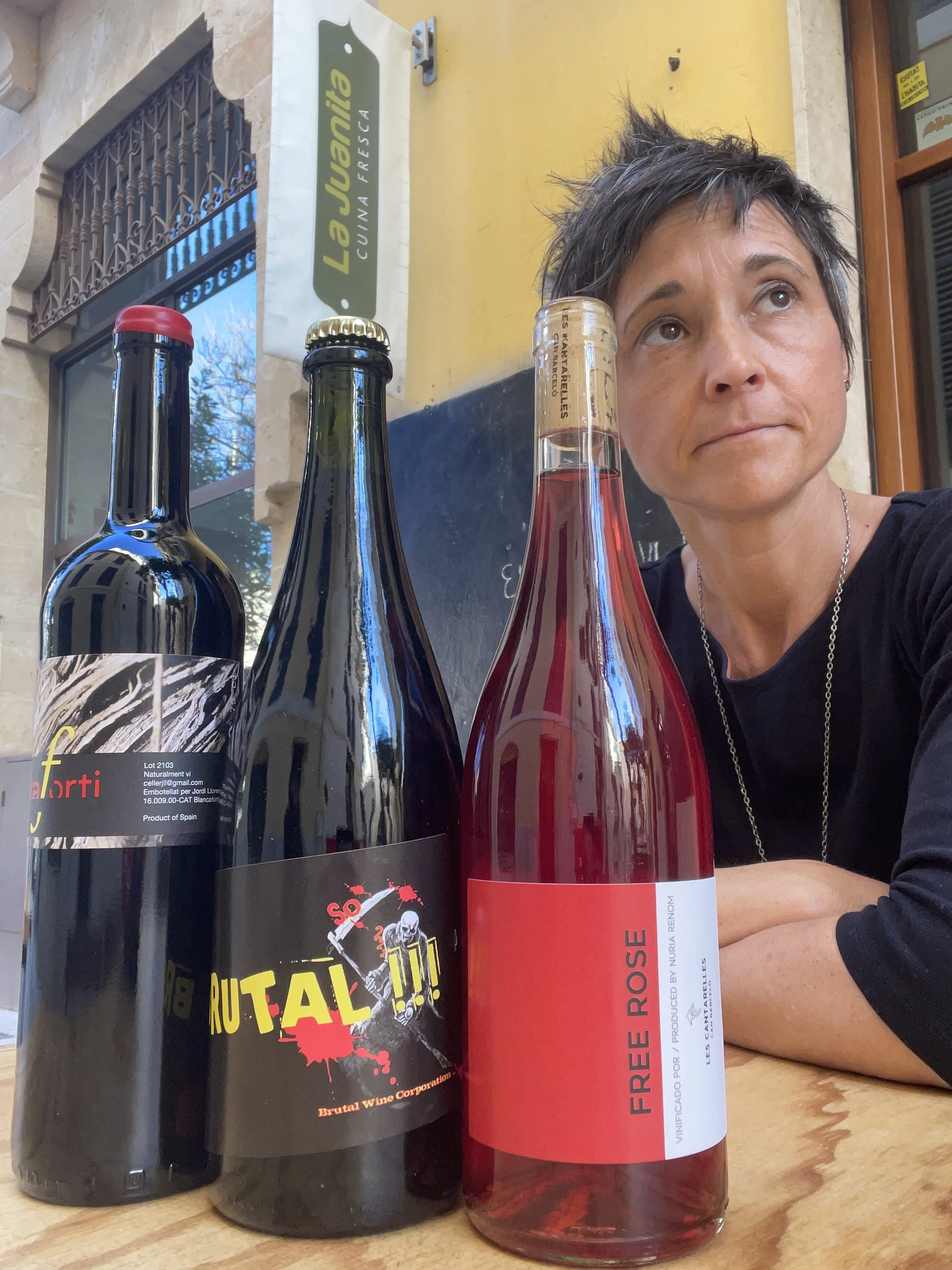 Three bottles of wine placed on a wooden table outside a restaurant called La Juani. A woman with short dark hair is sitting behind the bottles, with part of her face visible in the photo.