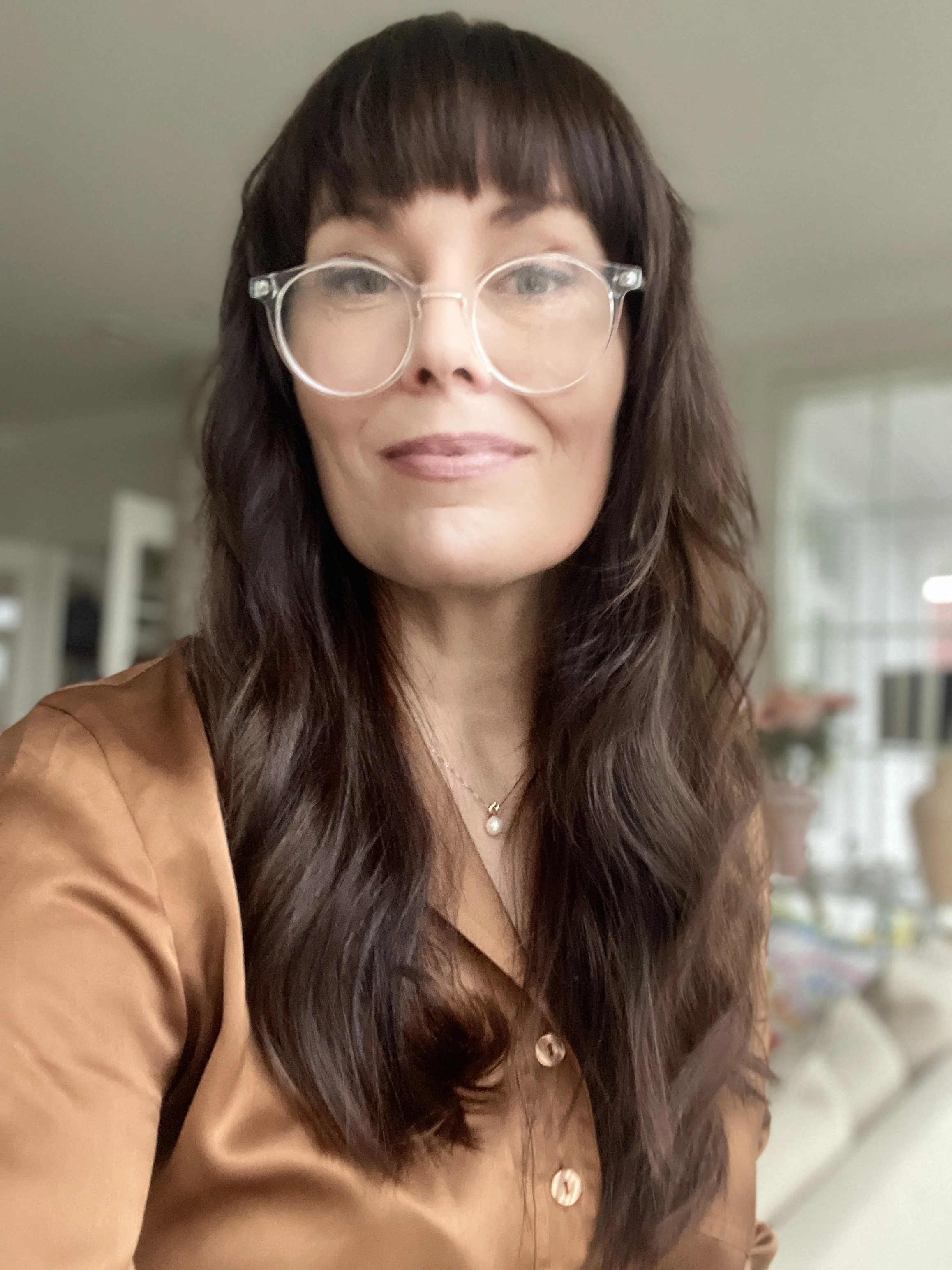 A woman with long wavy brown hair, wearing large clear glasses, a gold necklace with a pearl pendant, and a brown blouse, smiling softly, in a bright room with blurred background.