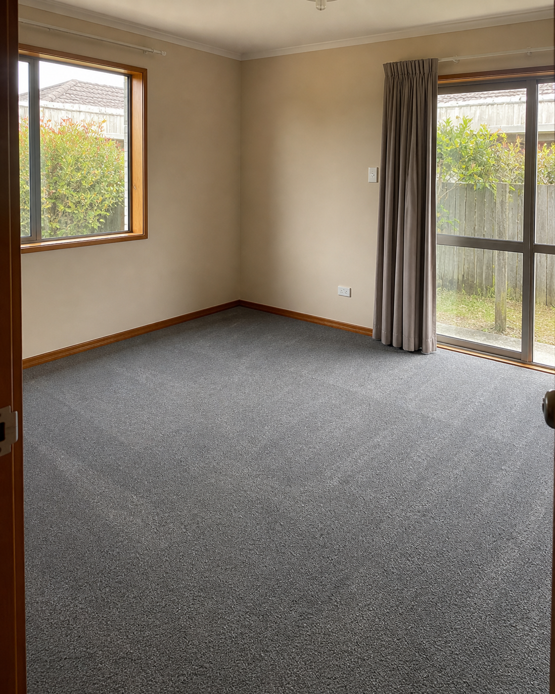 Empty room with beige walls, gray carpet, wooden trim, a window with wooden frame, and a glass sliding door with beige curtains, garden outside.