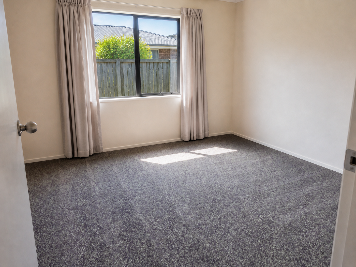 Empty bedroom with beige walls, gray carpet, and a large window with curtains, overlooking a backyard with a wooden fence and neighboring house.
