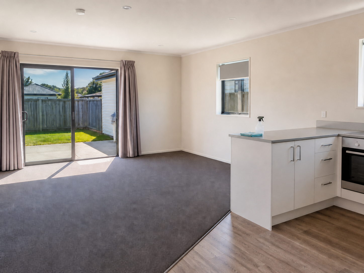 Empty living area with sliding glass door leading to backyard, small window, white cabinets, and partly visible kitchen with wood flooring.