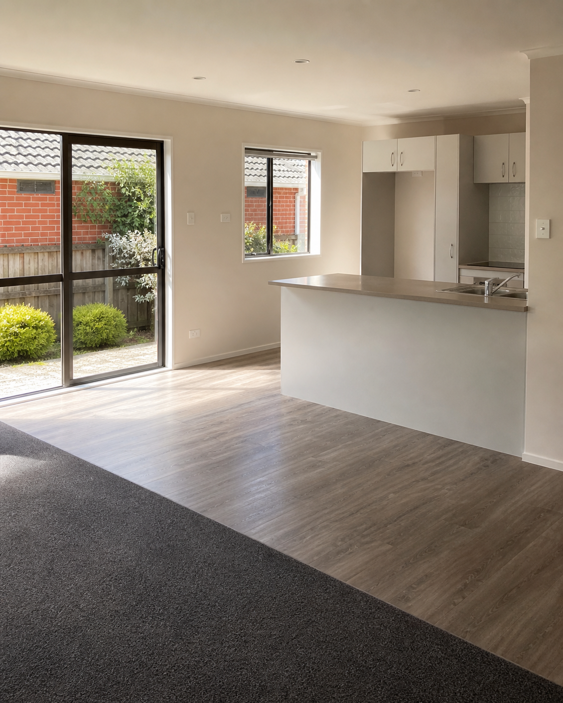 Empty living space with a kitchen area, featuring a sliding glass door opening to a backyard with bushes, and a window overlooking the backyard. The room has light-colored wood flooring and a partial wall with a kitchen counter.