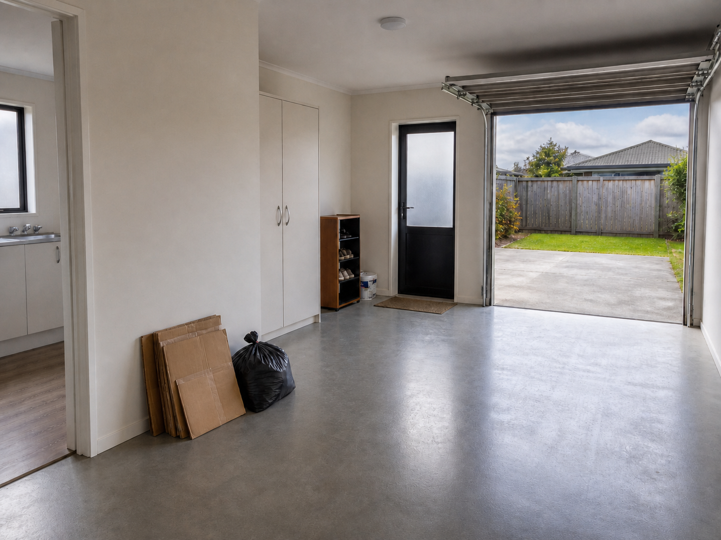 Empty garage with open door leading to backyard, containing cardboard, black trash bag, small shoe rack, cabinets, and a window.
