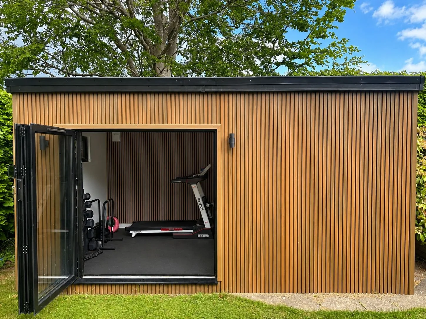 Modern wooden outdoor gym shed with open sliding glass door revealing workout equipment inside, including a treadmill, kettlebells, and dumbbells, set against a backdrop of trees and blue sky.