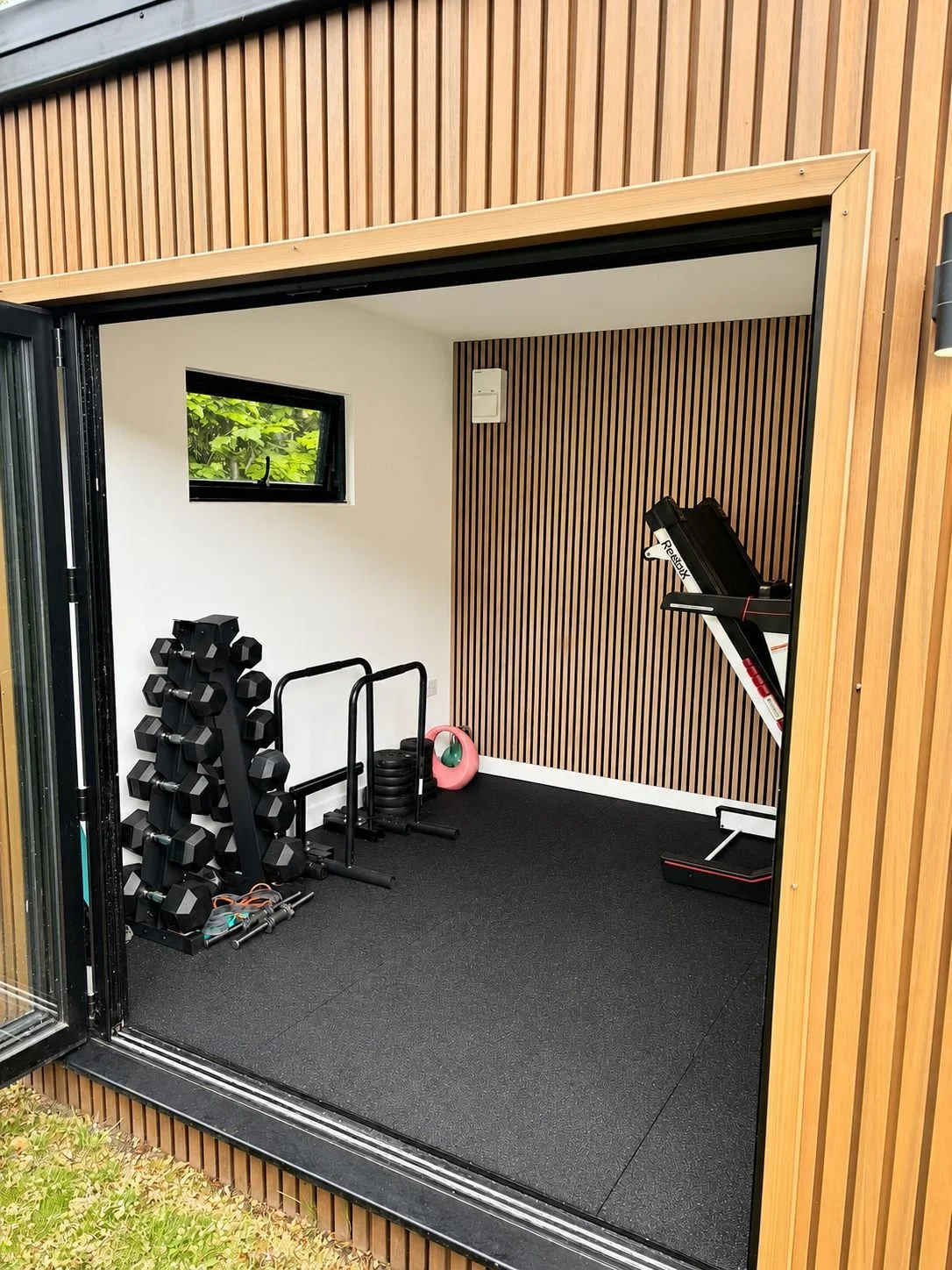 Small home gym with dumbbells, weight plates, resistance bands, pink kettlebell, and treadmill inside a wooden shed.