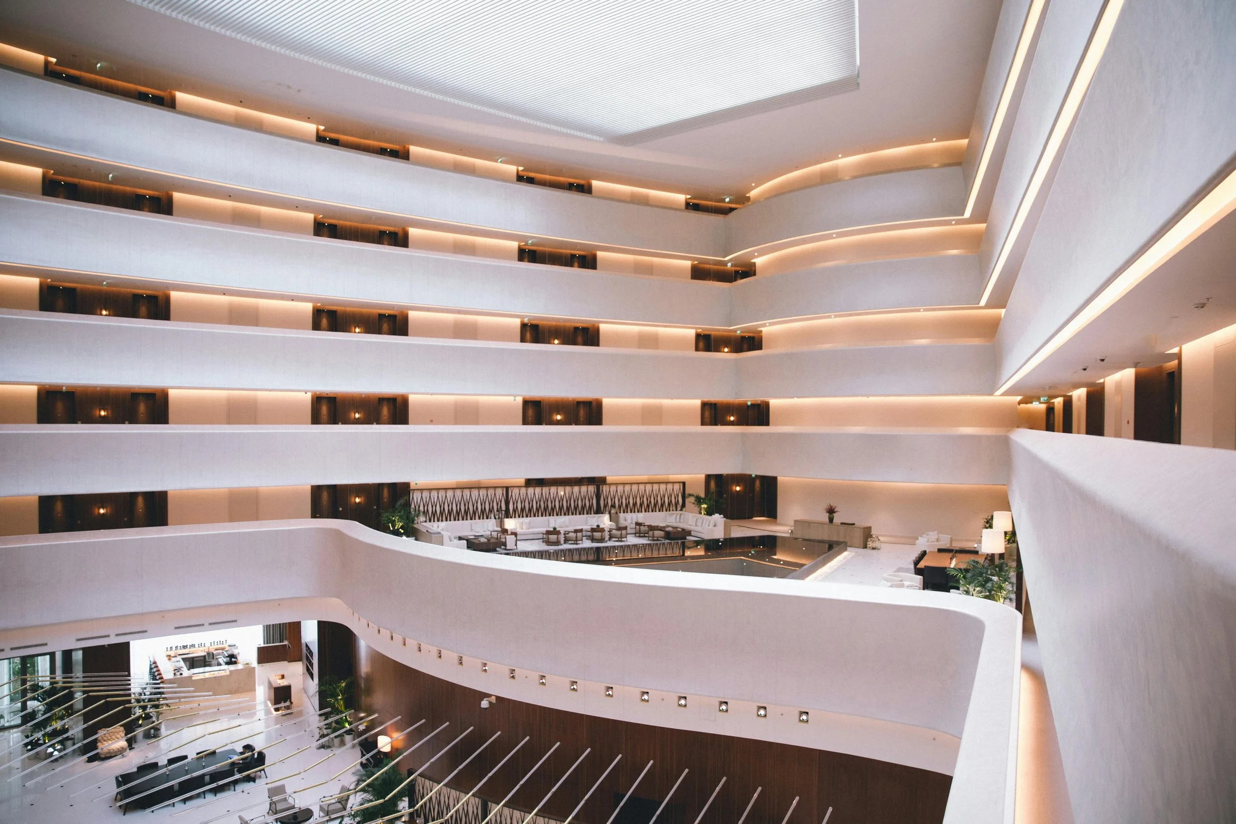 Hospitality - Interior of a modern hotel lobby with multiple floors, curved white balconies, warm lighting, and a seating area at the ground level.