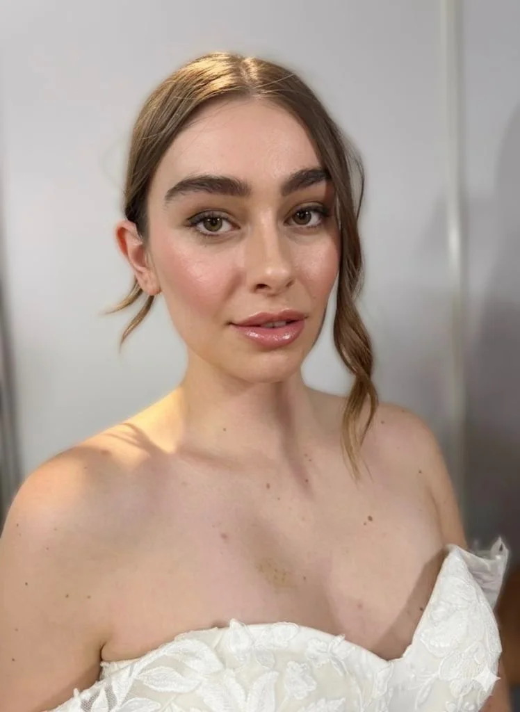 Close-up of a woman with light brown hair styled with a loose wave, wearing a strapless white dress with floral embroidery, standing in front of a plain wall.