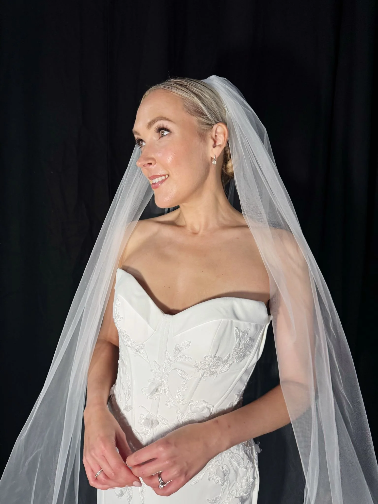 Close-up of a bride with blonde hair and earrings, wearing a white wedding dress with floral embroidery, and a veil, standing against a black backdrop.