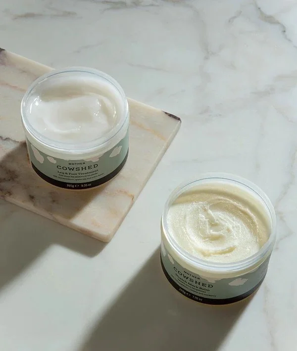 Two open jars of Mother Cow Shed body butter on a white marble surface, with one jar placed on a small marble slab.